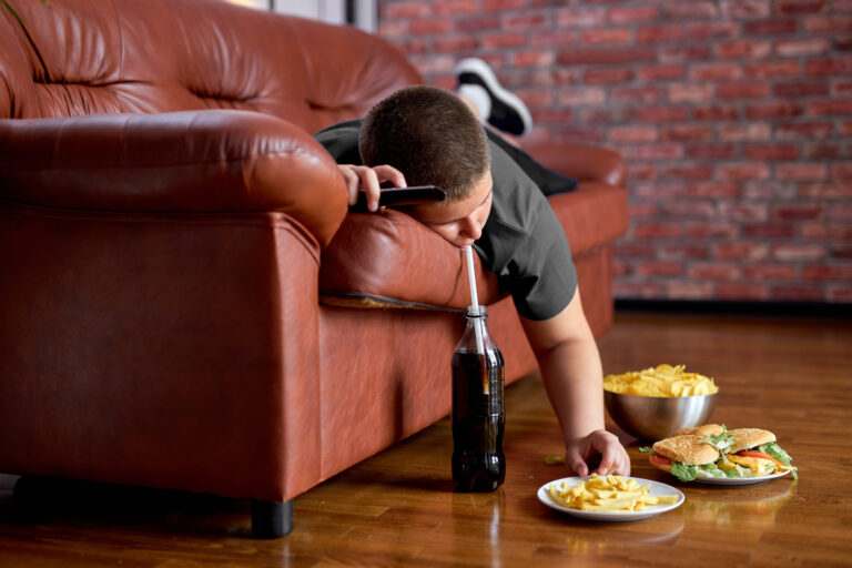 Man lying on couch eating fries and drinking beer, symbolizing sedentary habits and poor diet contributing to inflammation and depression.