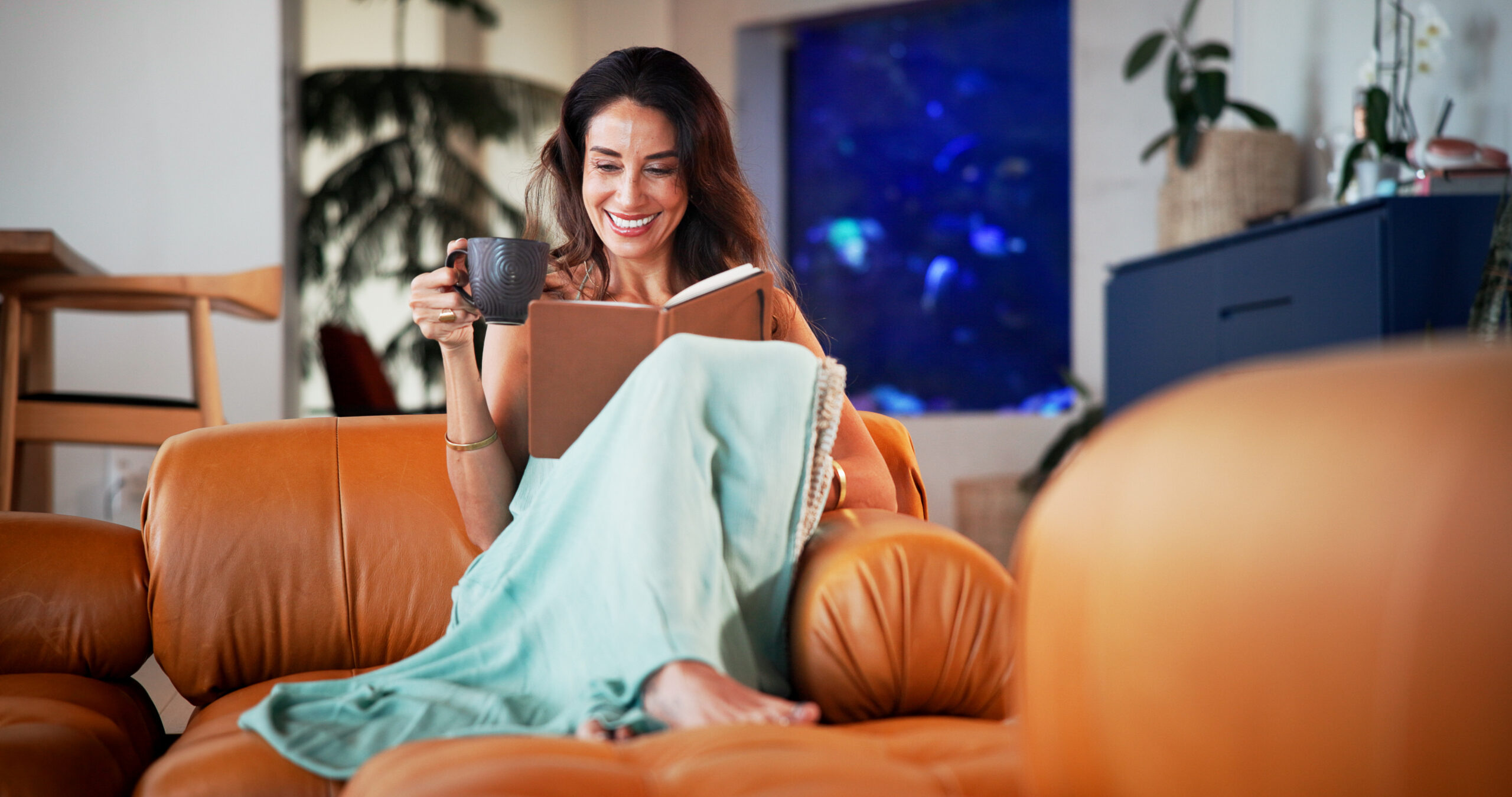 A woman relaxes peacefully on her couch with a book and coffee — representing accessible PTSD telehealth treatment from home in Virginia